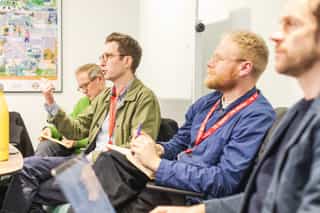 An image of three men in a row, one man is talking out while the others listen on