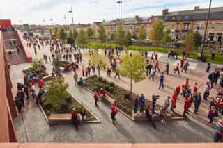 An high angle, exterior shot of Anfield entrance, showing the planting and seating while fans walk past and some use the seating