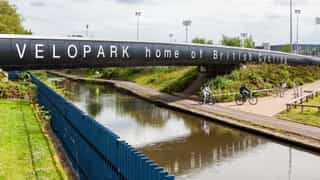 An image of a bridge over a canal with the words 'VELOPARK home of British Cycling' on the side, two people are cycling down the path on the canal