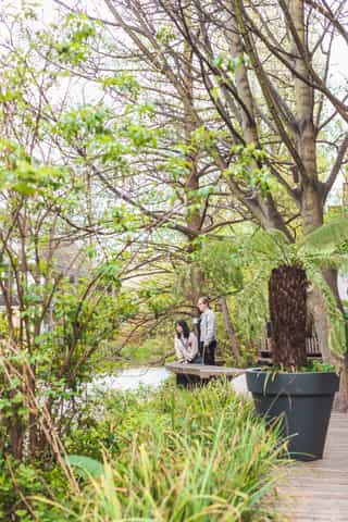 An image of two women looking down at water on the edge of a canal, green trees and planting surround the space