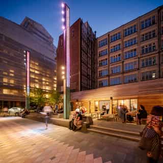 After shot of where a back alley used to sit in Manchester, now there is a hand built timber pub called the pilcrow, seating steps and a tall illuminated lighting totem.