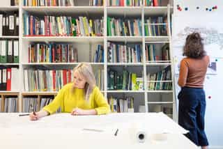 An image of a woman sat writing, with a wall of books behind her