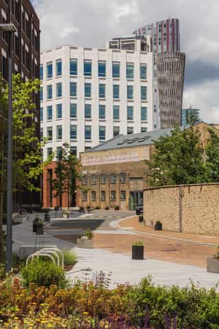 An image of the pathways around Sugar House Island, with bike racks, seating and planting down either side. The Pathways bend round towards buildings in the distance