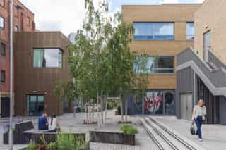An exterior shot of a courtyard at Sugar House Island, two people are using the seating and chatting, while a woman walks by on the phone, with large birch trees in the middle of the space